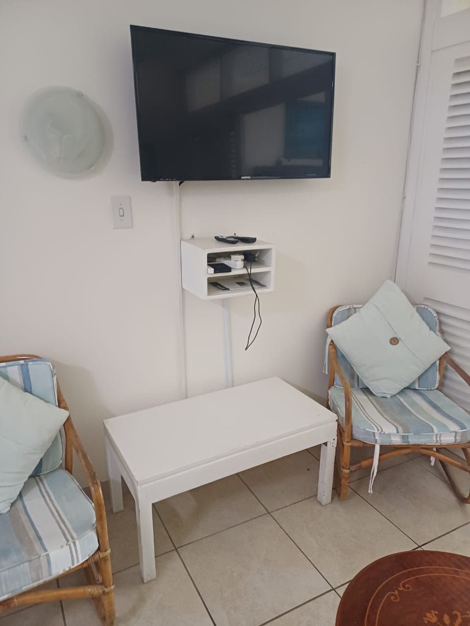 A wall-mounted TV above a small white shelf, a white coffee table and two cane chairs with blue and white striped cushions